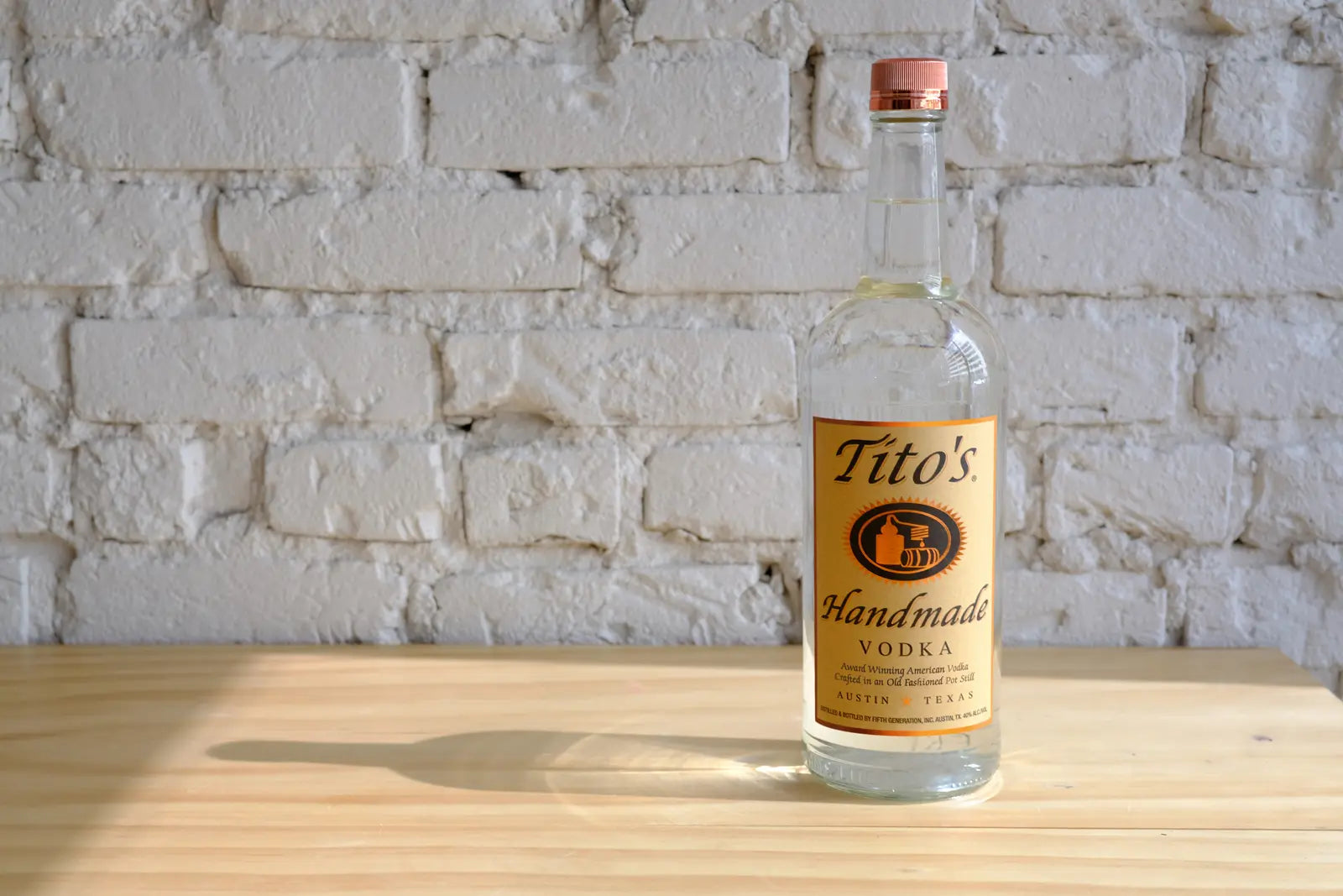 Bottle of Tito's Handmade Vodka on a wooden table against a white brick backdrop.