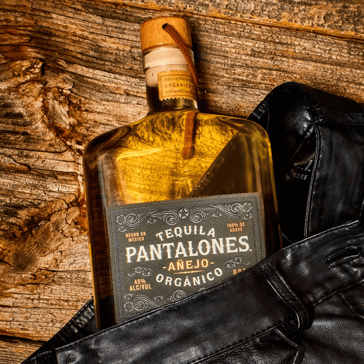 Pantalones Añejo Tequila bottle resting on leather pants against a rustic wooden background.