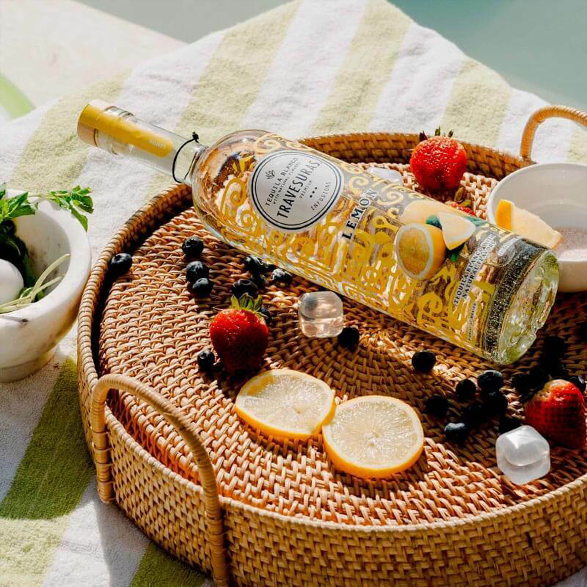 Travesuras Lemon Tequila bottle on a woven tray with fresh strawberries and lemons for a refreshing sip.