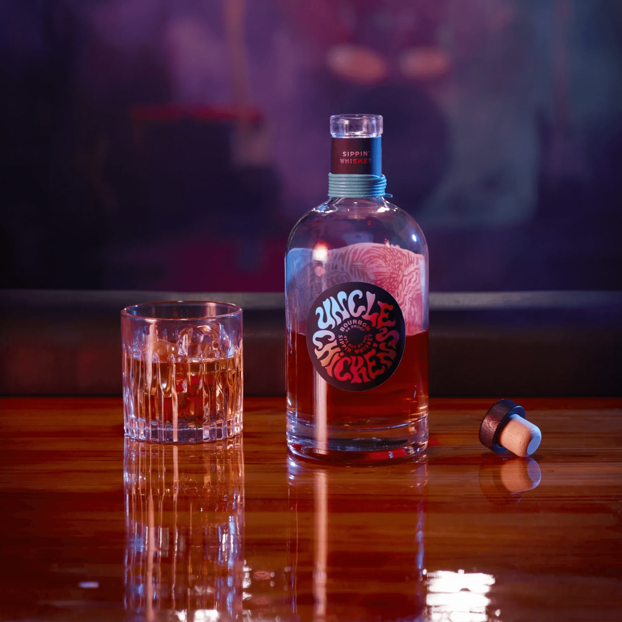 Uncle Chicken's Sippin' Whiskey Bourbon bottle next to a glass of whiskey on a wooden table.
