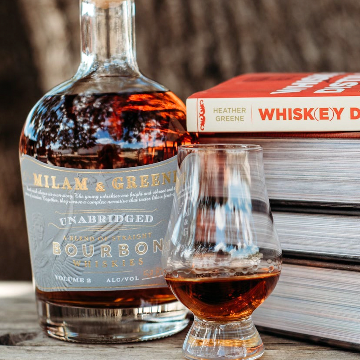 Milam and Greene Unabridged Bourbon Vol 2 bottle with glass, surrounded by whiskey books on a wooden surface.