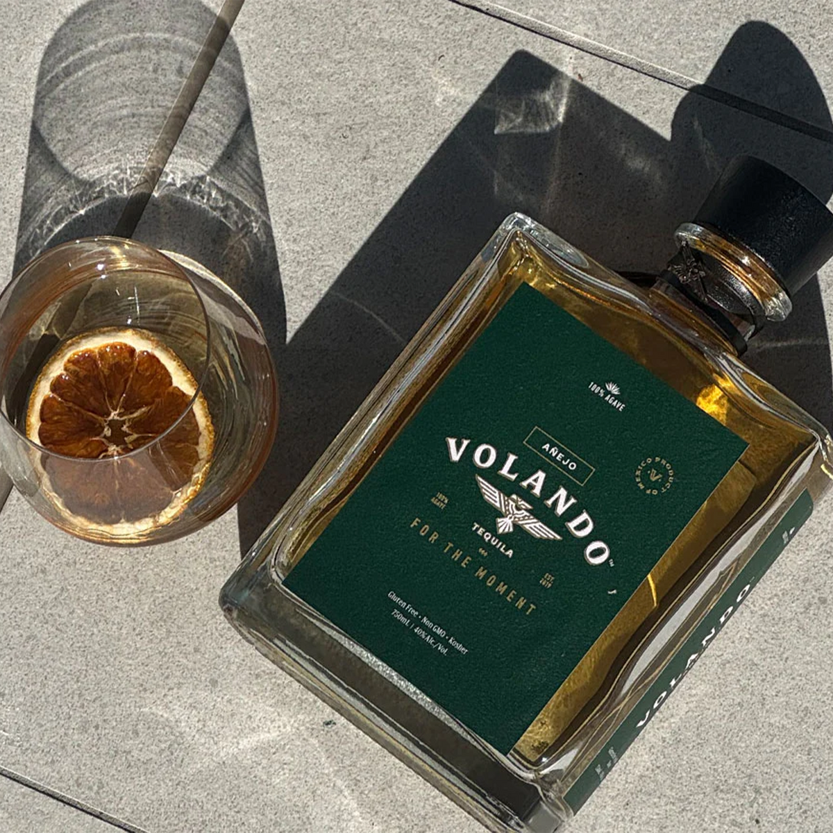 Bottle of Volando Añejo Tequila with a glass and dried orange on a stone surface.