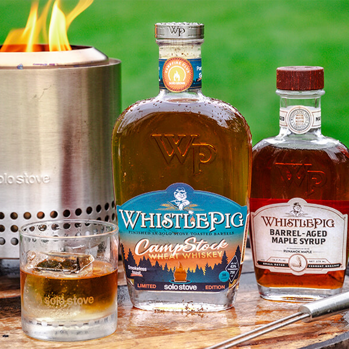 WhistlePig CampStock Wheat Whiskey bottle next to a glass of whiskey, campfire, and barrel-aged maple syrup.