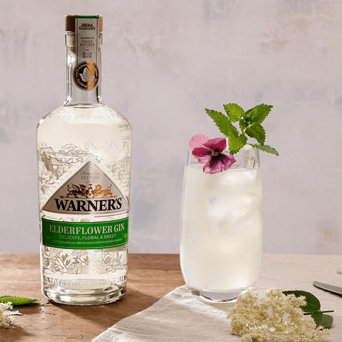 Warner's Elderflower Gin bottle with a refreshing elderflower cocktail garnished with mint and flower.
