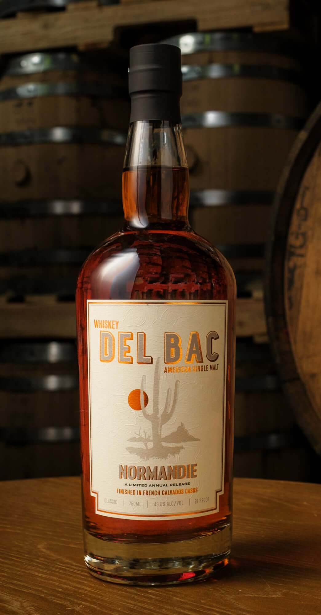 Del Bac Normandie Limited Release Whiskey in front of wooden barrels, featuring its signature label and rich amber color.