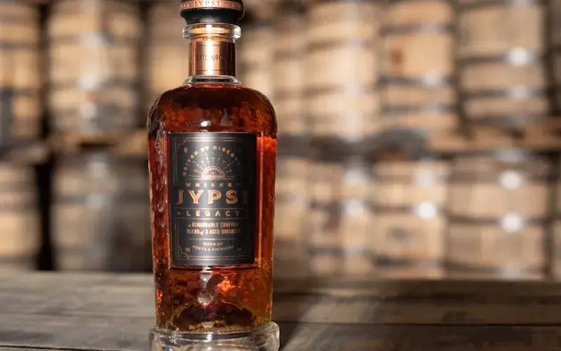 Bottle of Whiskey JYPSI Legacy Batch 001 "The Journey" set against a backdrop of whiskey barrels.