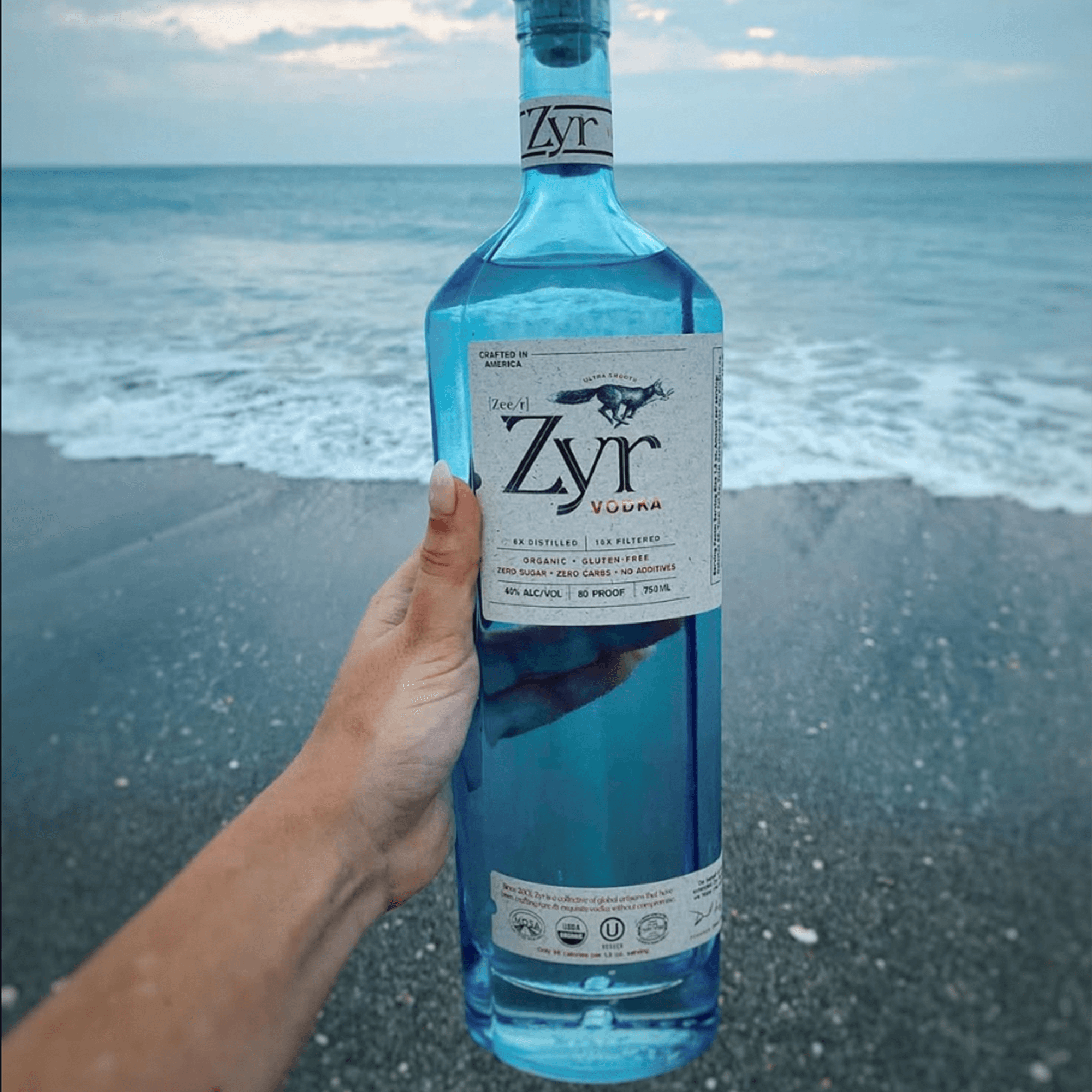 Zyr Organic Vodka bottle held by a hand, with ocean waves in the background, showcasing its vibrant blue packaging.