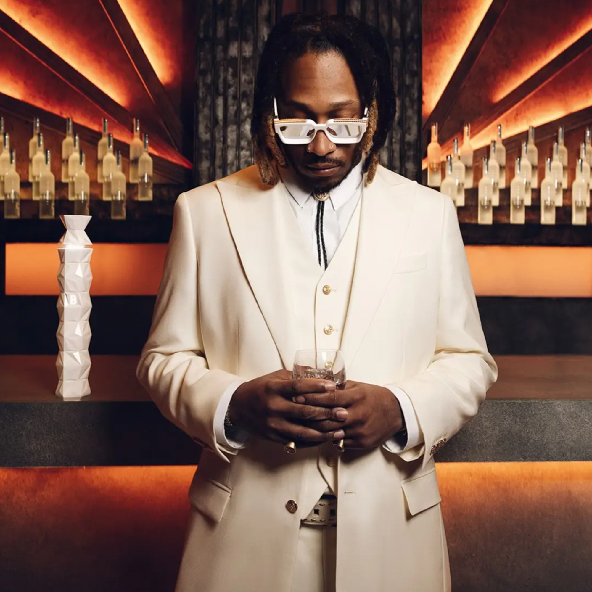 Elegantly dressed man in a white suit holding a glass, with a bar backdrop highlighting luxury vodka bottles.