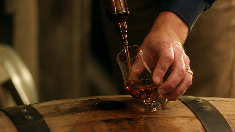 Pouring Henry McKenna Single Barrel Bourbon into a glass from a wooden barrel.