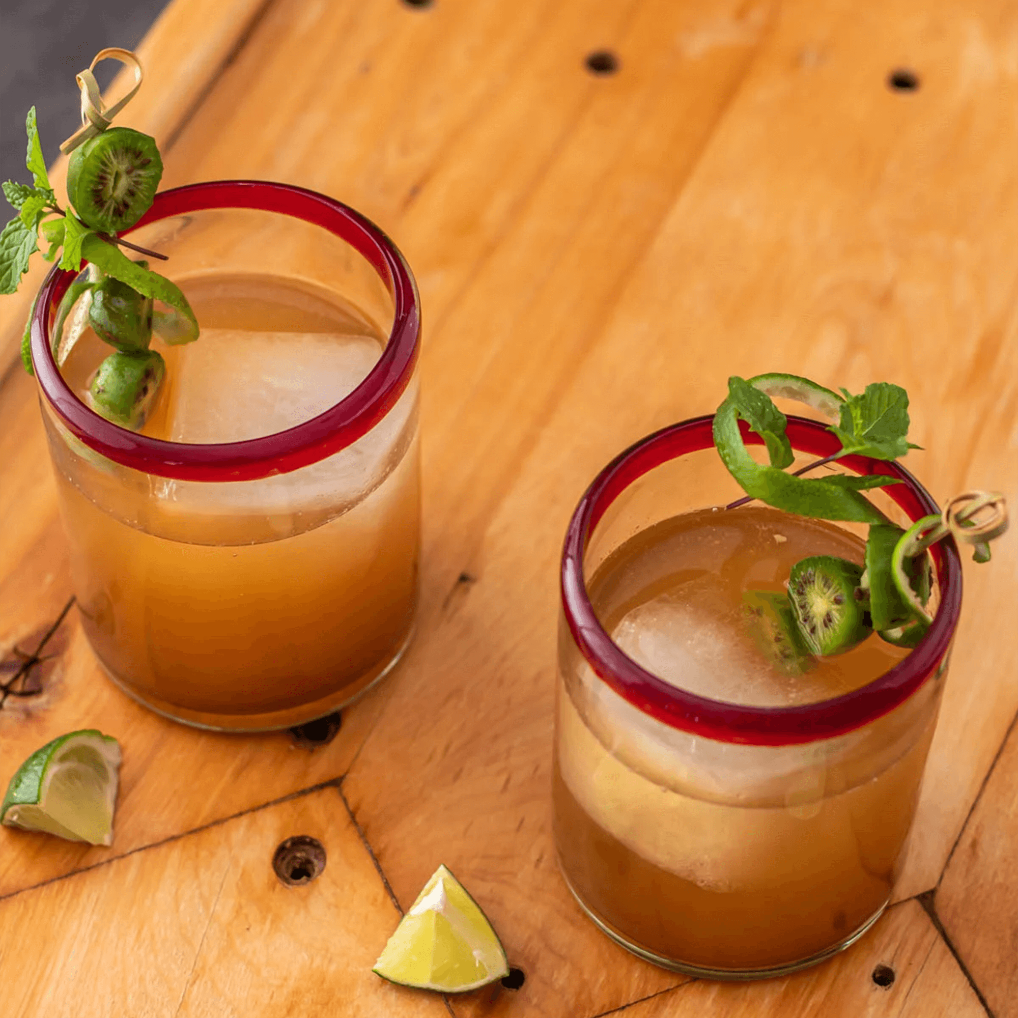 Two cocktails garnished with herbs and lime on a wooden surface, perfect for enjoying Casa Silencio Mezcal.