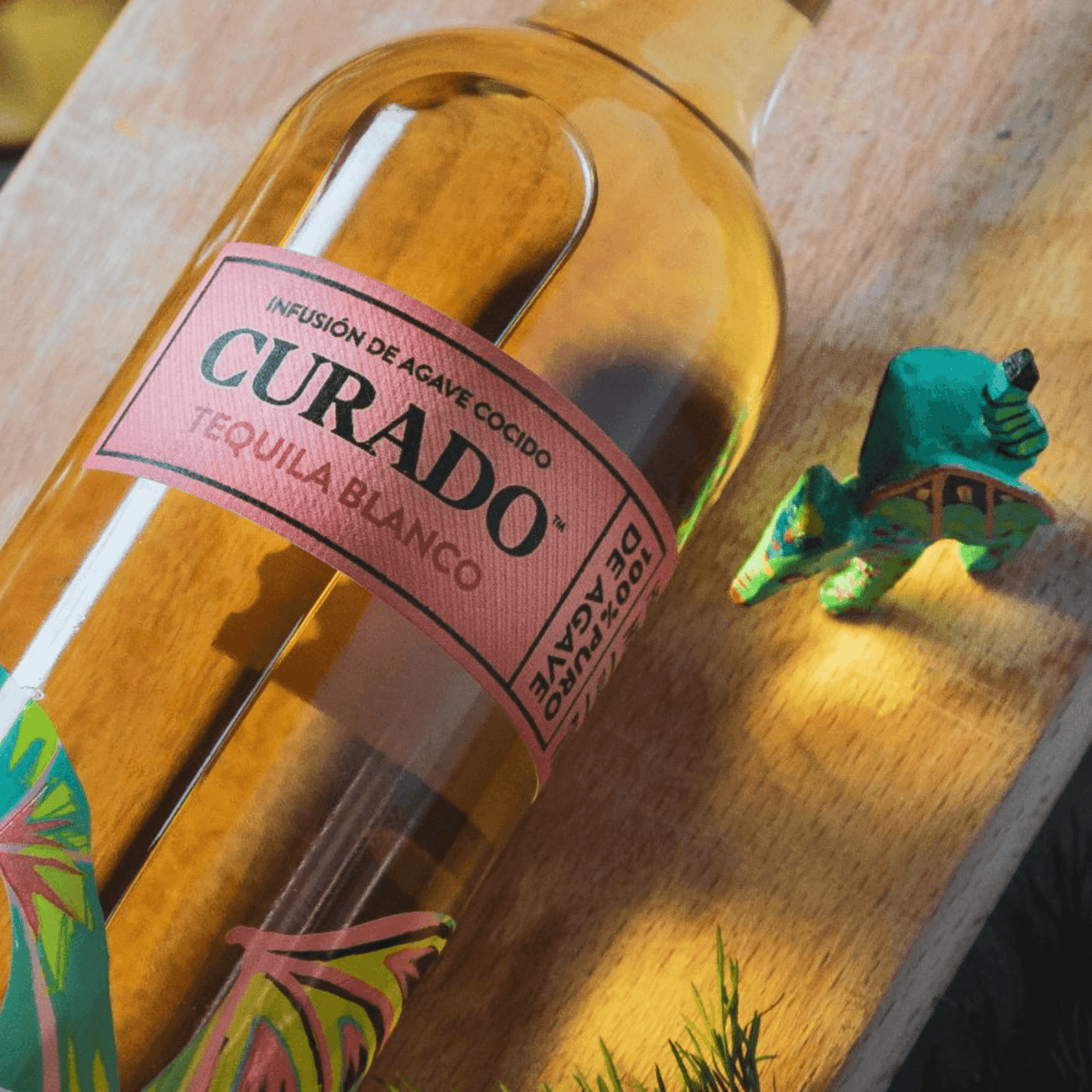 Curado Blue Agave Blanco Tequila bottle with vibrant floral design and a Tejón figurine on wood surface.