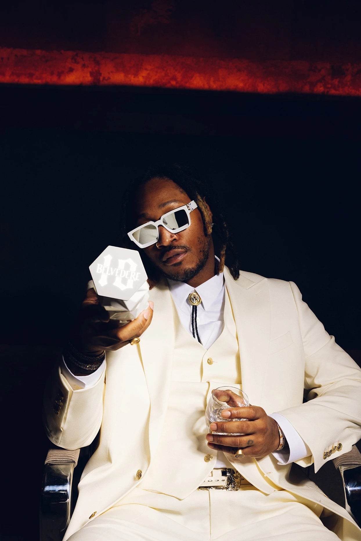 Stylish man in a white suit holding a geometric object, sipping a drink in a dimly lit venue.