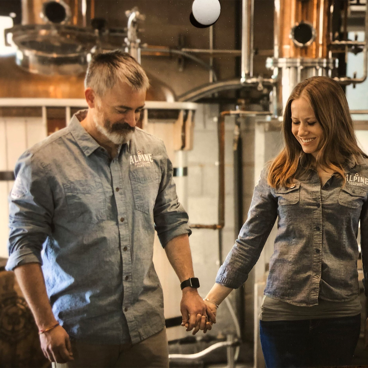Couple holding hands in front of distilling equipment, wearing Alpine Distilling shirts, showcasing craftsmanship.