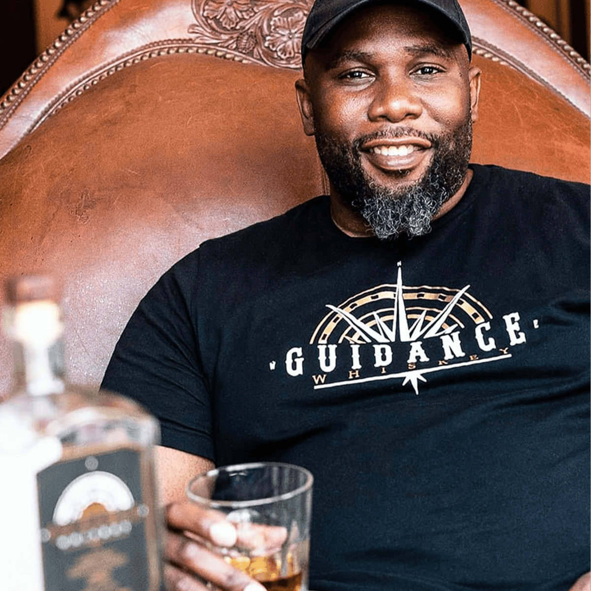 Smiling man holding a glass of whiskey, wearing a Guidance Whiskey t-shirt, seated in a leather chair.
