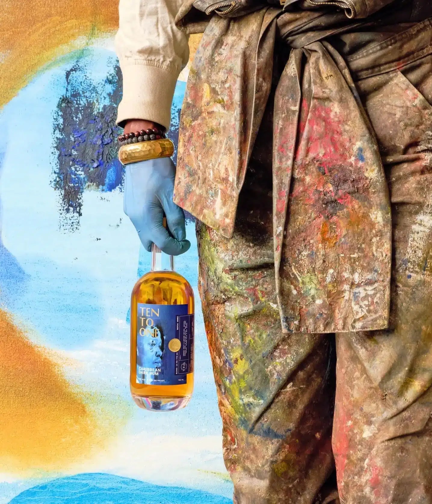 Artist holding Ten To One Devin B. Johnson Caribbean Dark Rum bottle in front of colorful artwork.