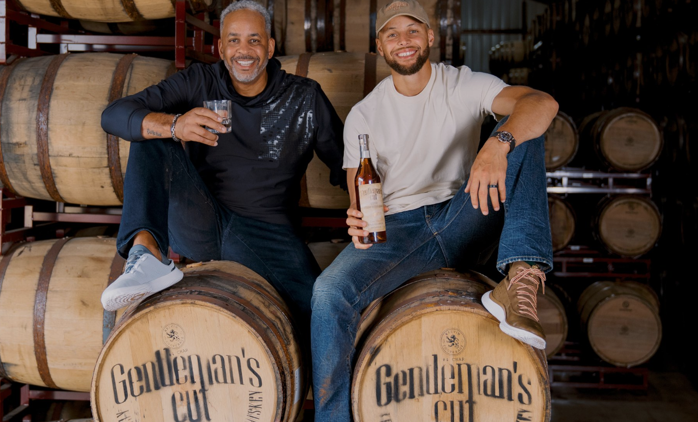 Two men enjoying Gentleman’s Cut Bourbon on whiskey barrels at Game Changer Distillery, celebrating the new spirits launch.