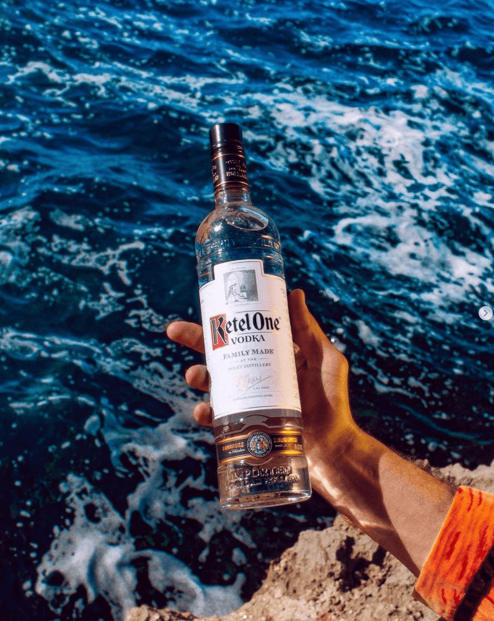 A hand holding a bottle of Ketel One Vodka by the ocean, showcasing its crisp, fresh appeal.