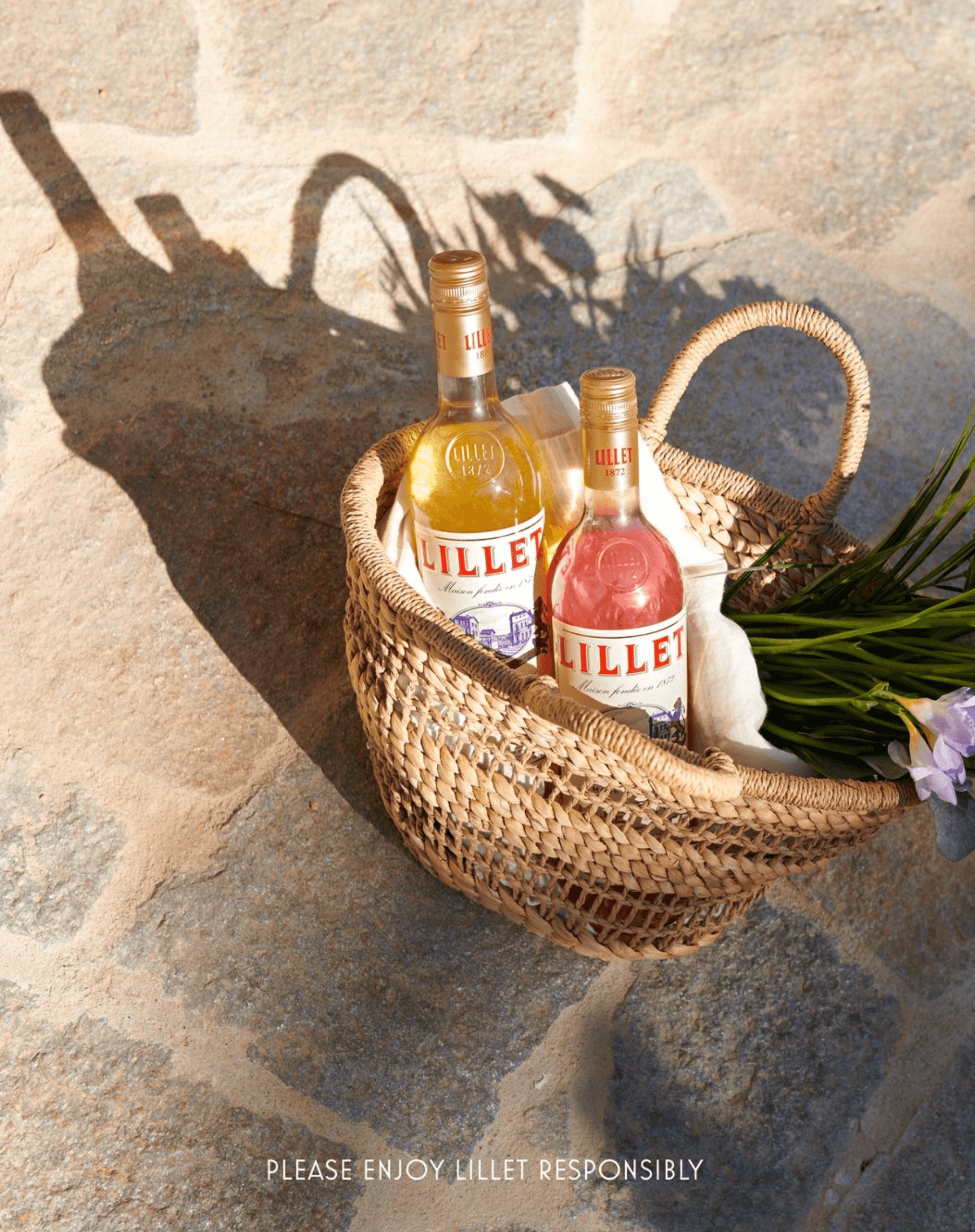 Two bottles of Lillet aperitif in a woven basket with flowers, set on a stone surface.