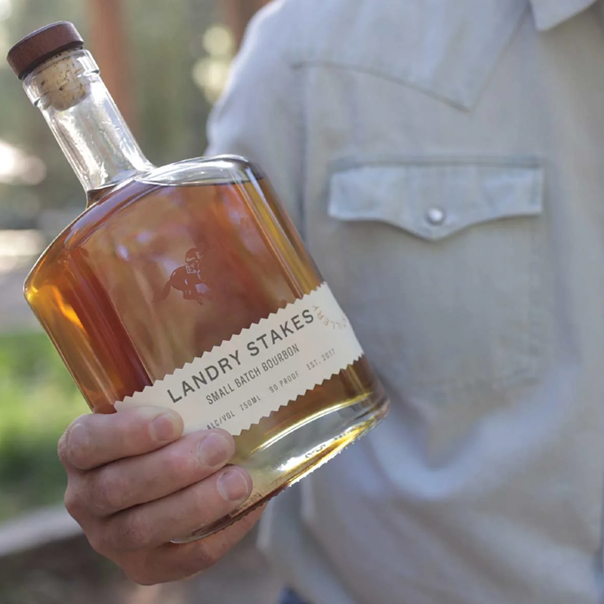 Bottle of Landry Stakes Small Batch Bourbon Whiskey held outdoors, showcasing its bright amber hue.