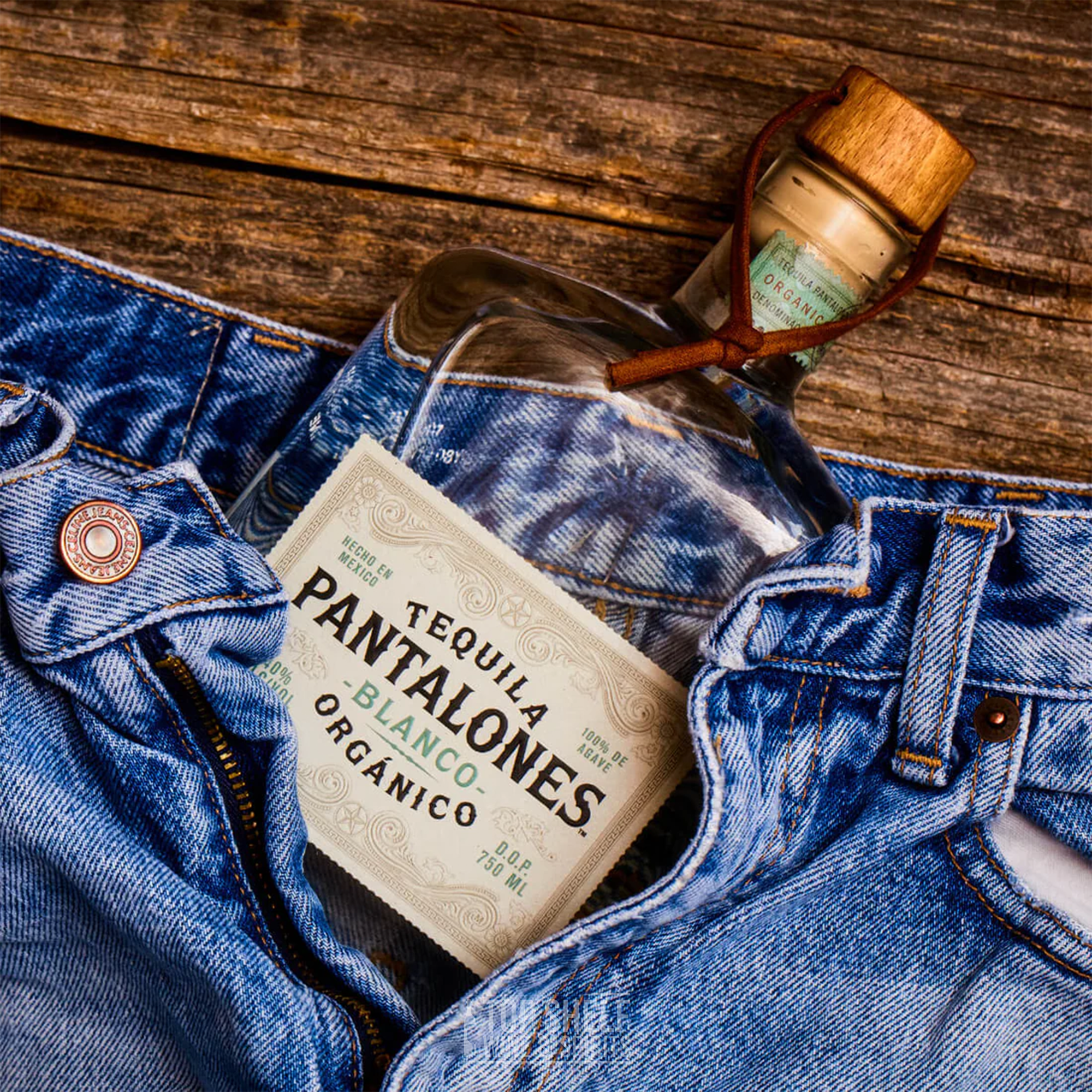 Bottle of Pantalones Blanco Tequila nestled in denim jeans, showcasing organic tequila on rustic wooden background.