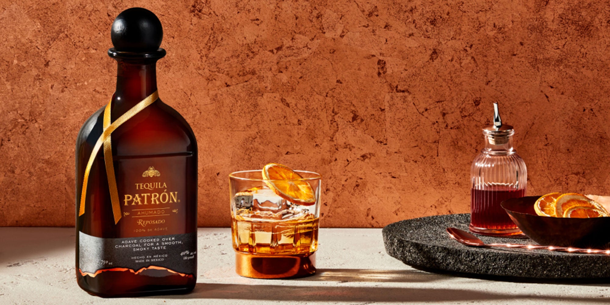 Bottle of Patrón Ahumado Reposado tequila with a glass, garnished with orange, showcasing its smooth and smoky flavor profile.