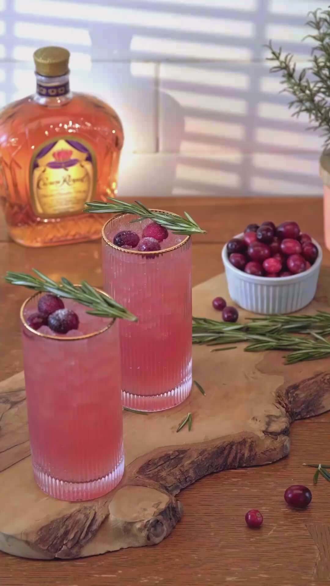 Two pink cocktails garnished with rosemary and cranberries, with a bottle of Crown Royal whisky in the background.