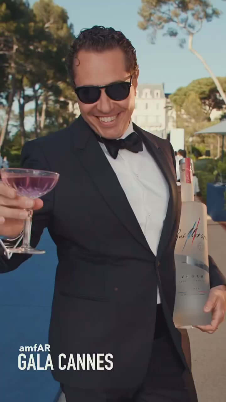 A man in sunglasses holding a cocktail at the amfAR Gala in Cannes, with a bottle of premium vodka in the background.