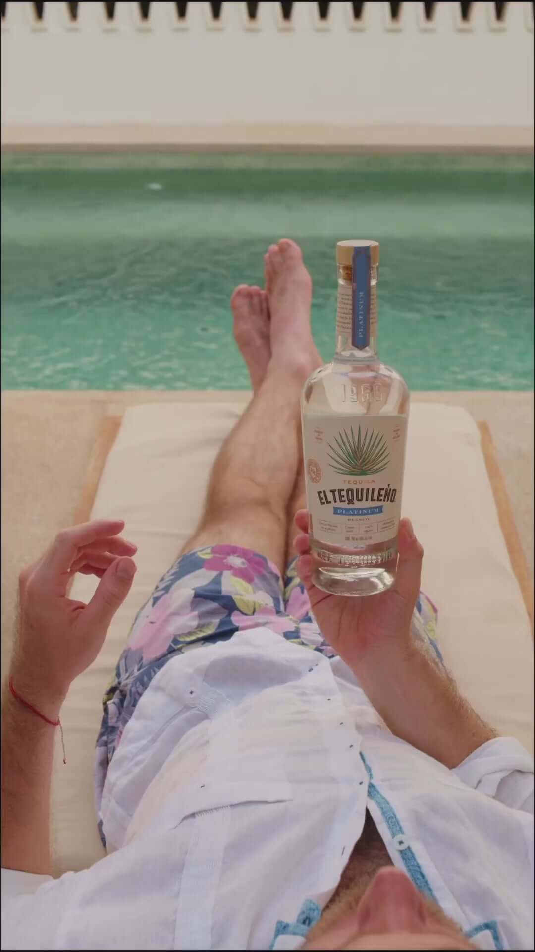 Person relaxing by the pool holding a bottle of El Tequileño Platinum Blanco Tequila, perfect for summer cocktails.