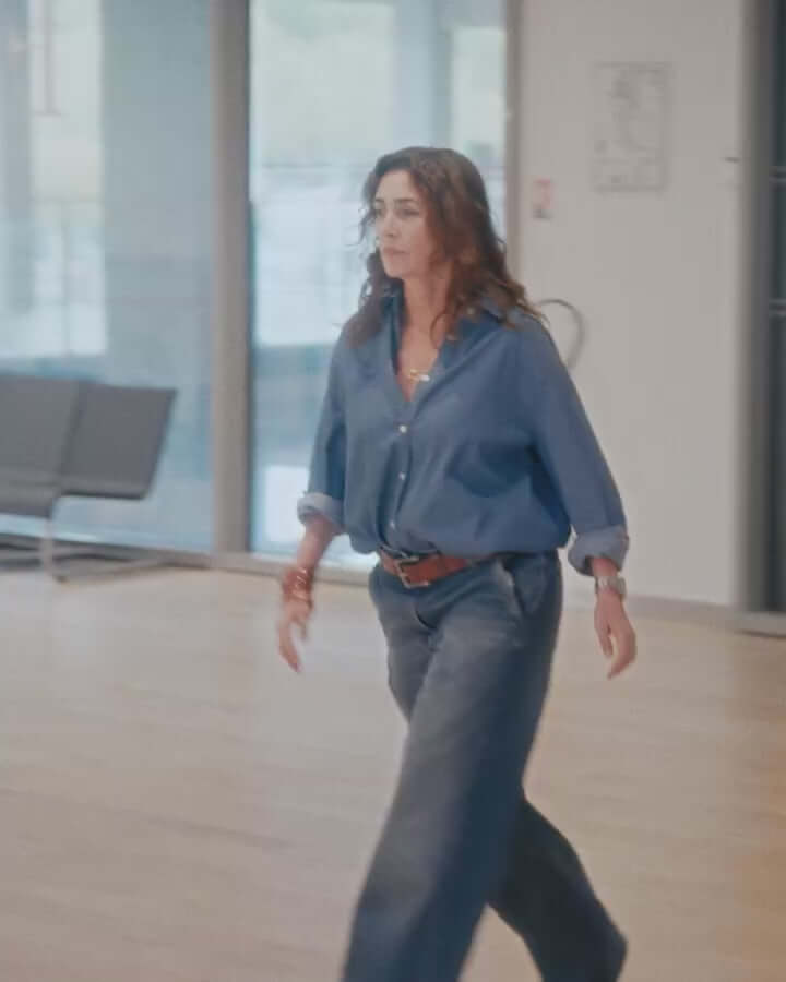 Woman walking in a casual outfit featuring a blue shirt and wide-leg jeans in a modern interior space.