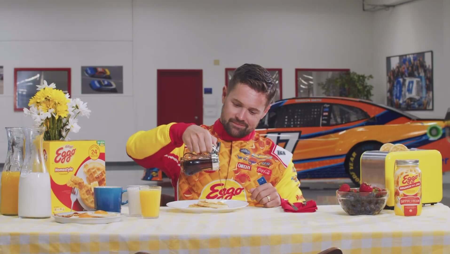 Man in Eggo jersey pours syrup over waffles at a festive breakfast table with cars in the background.