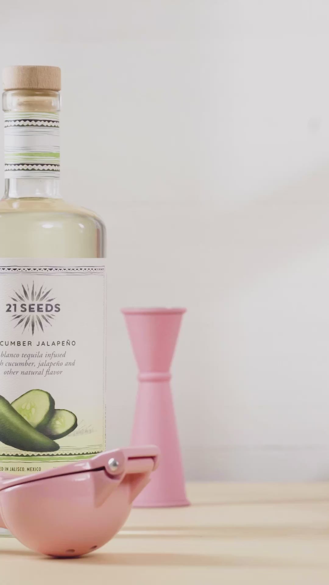 21 Seeds Cucumber Jalapeño Tequila bottle on a table next to a pink jigger and a lime slicer, highlighting its all-natural ingredients.