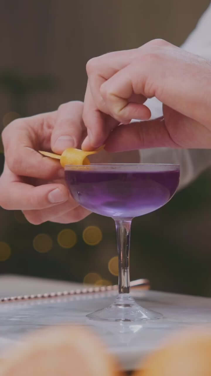 Hand garnishing a purple cocktail with a lemon twist, showcasing Empress 1908 Gin in an elegant glass.