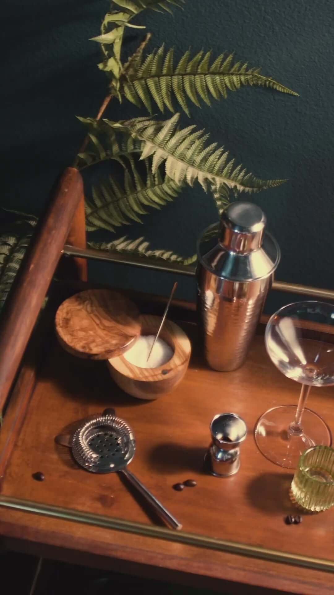 Cocktail preparation setup with a shaker, strainer, bowls, and foliage on a wooden tray.