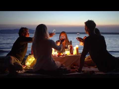 Group of friends toasting at a beach dinner with candles and drinks at sunset.