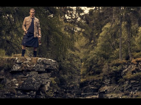 Man in a kilt standing on a rocky ledge in a Scottish landscape, holding The Sassenach blended Scotch whisky.
