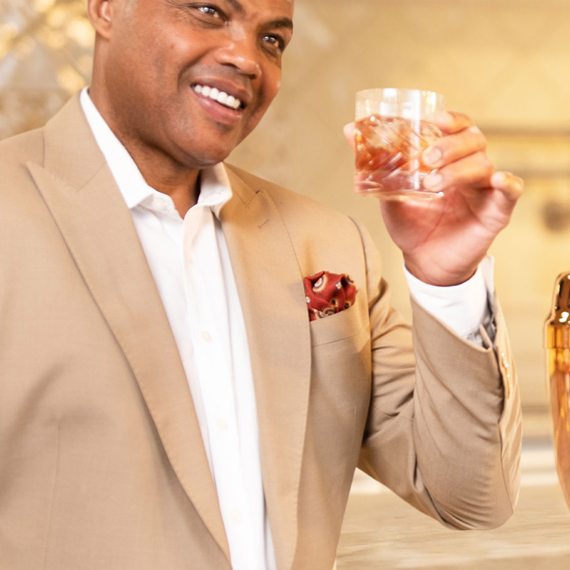 Man in beige suit holding a glass with a drink, smiling in a stylish bar setting.