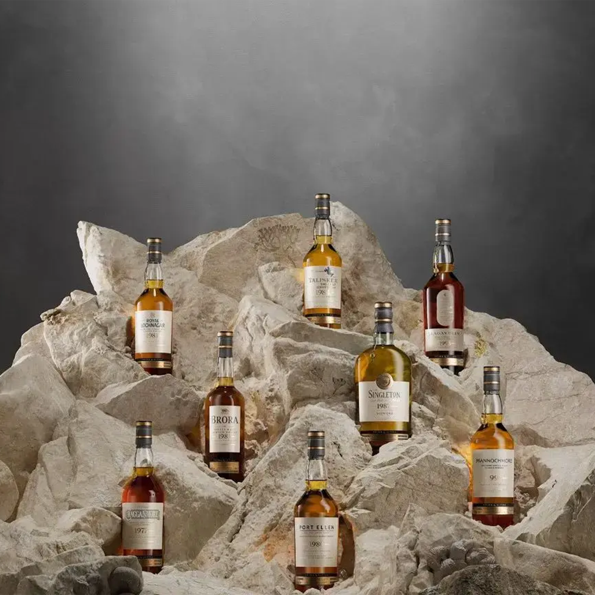 Display of seven premium Scotch whiskey bottles on a rocky backdrop, showcasing their rich golden hues.