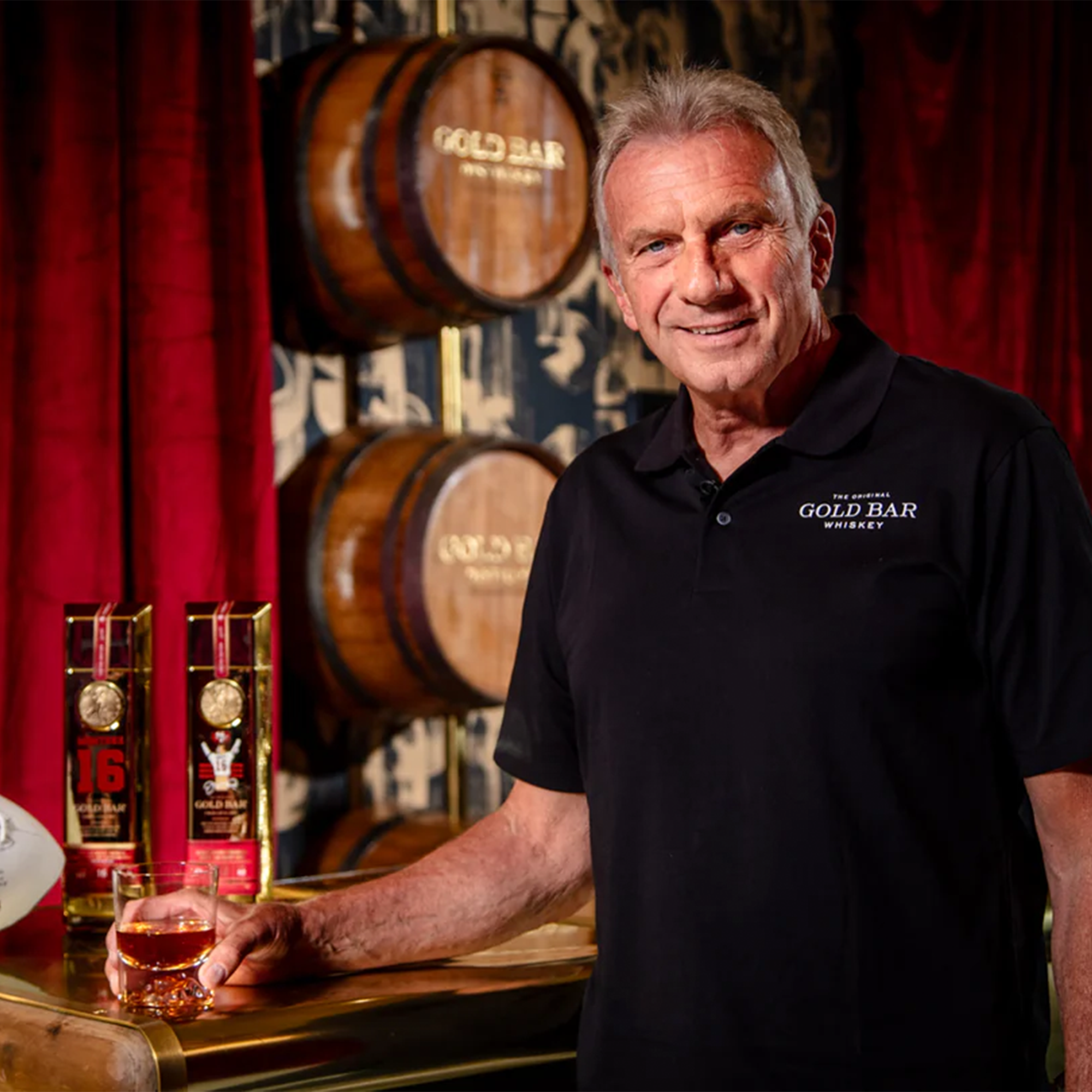 Joe Montana promoting Gold Bar Whiskey with awards, whiskey barrels, and a celebratory glass in a sophisticated setting.