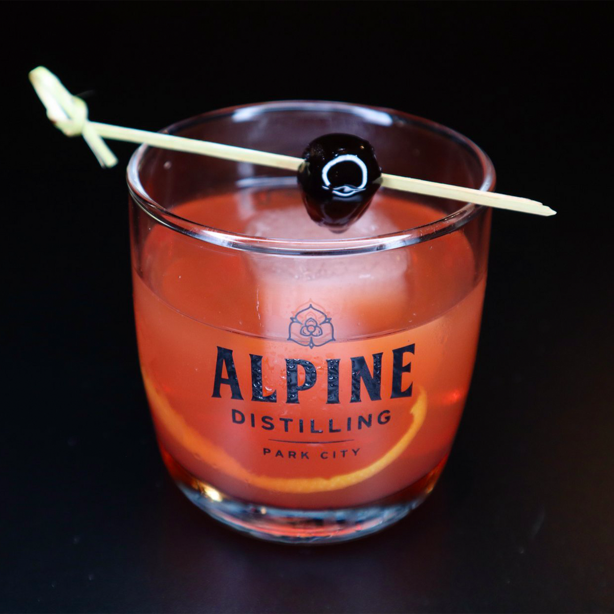 Cocktail served in an Alpine Distilling glass with cherry garnish and orange slice, reflecting the brand's signature style.