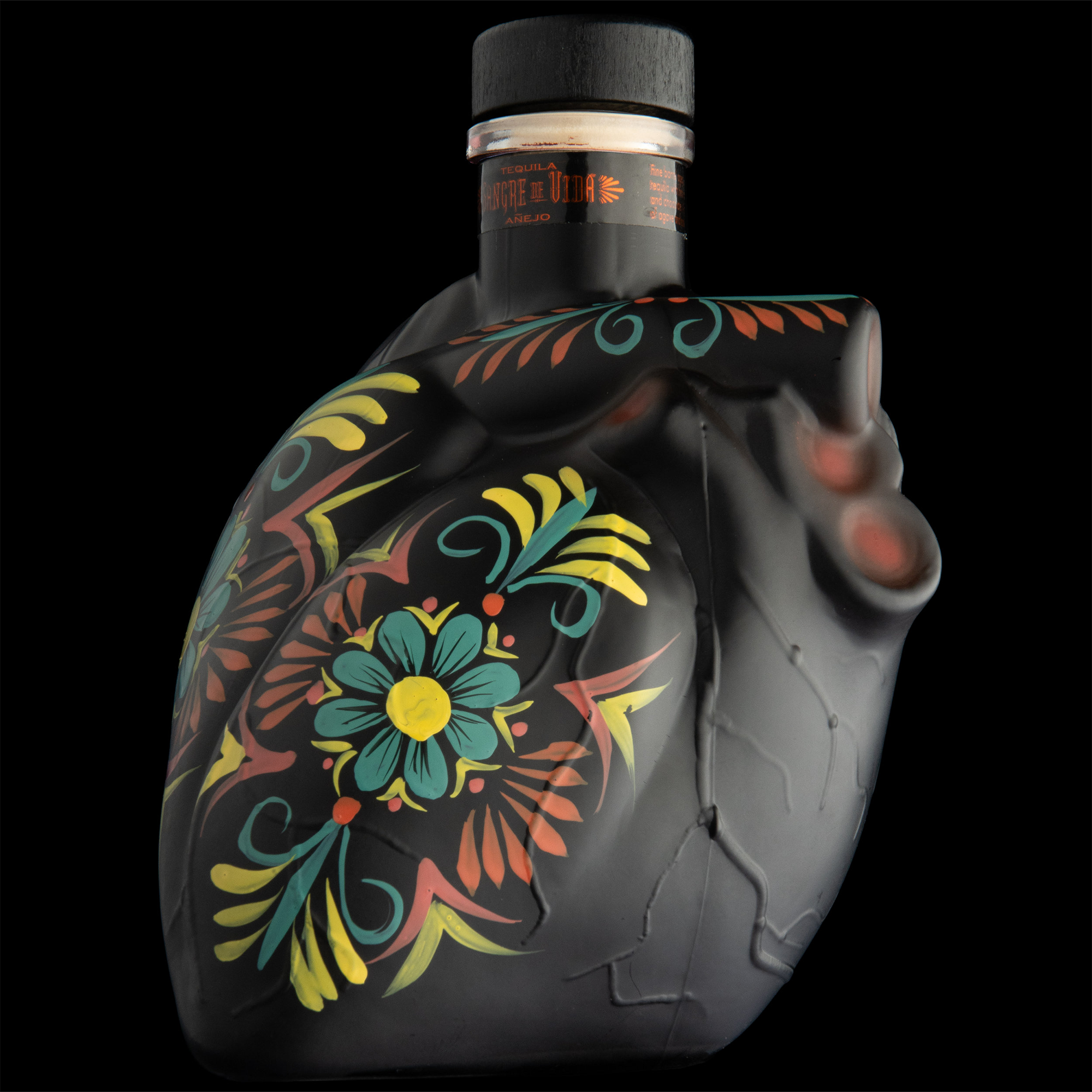 Sangre De Vida Añejo Tequila bottle designed like a heart, featuring colorful floral patterns on a dark background.