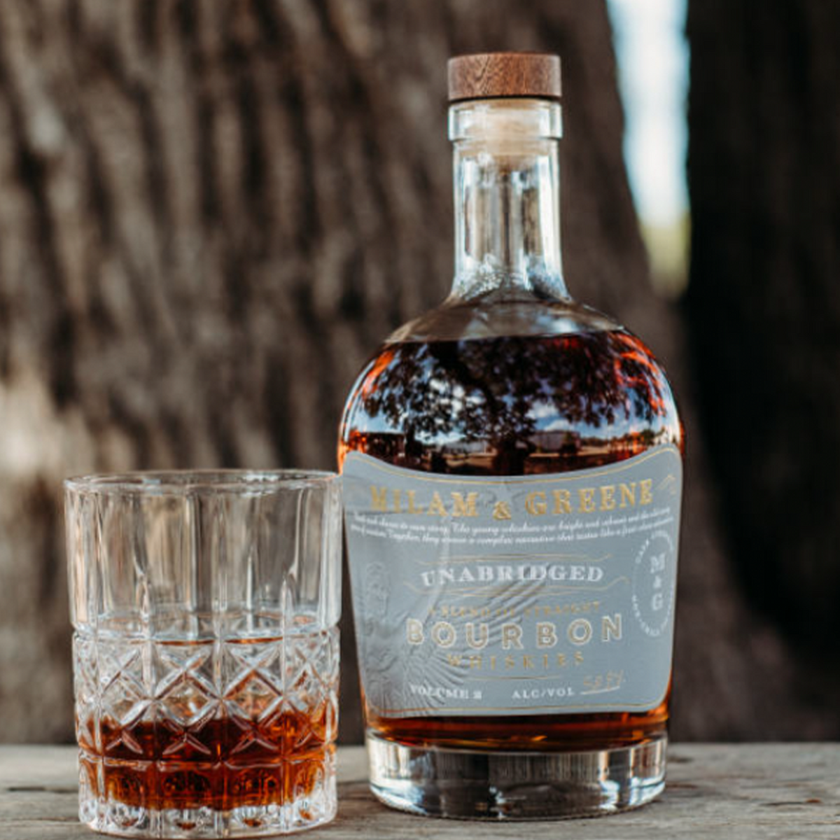 Milam and Greene Unabridged Bourbon Vol 2 in a bottle next to a glass of bourbon, showcasing its rich amber color.