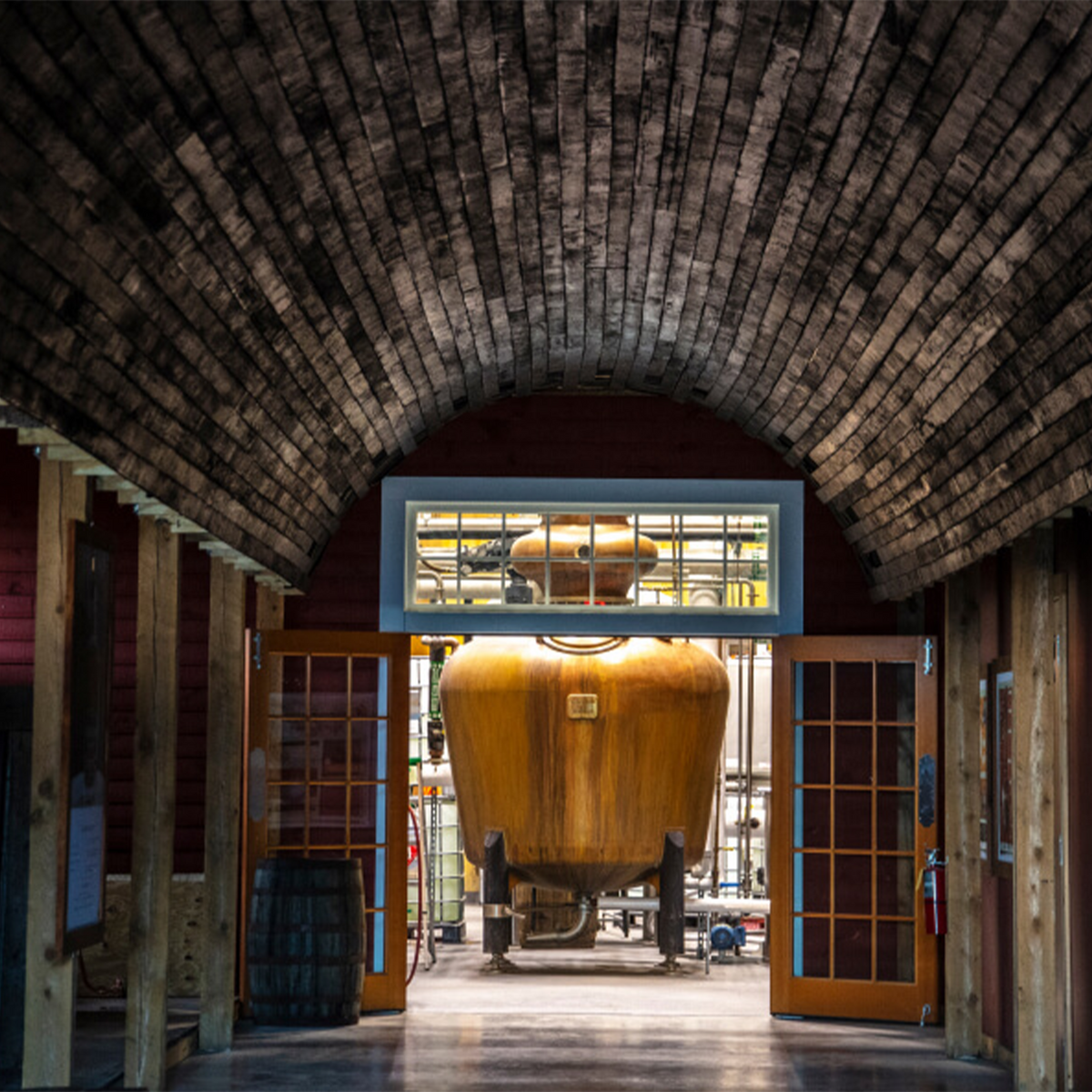 View of whiskey distillery hallway leading to a large wooden still under a textured ceiling.