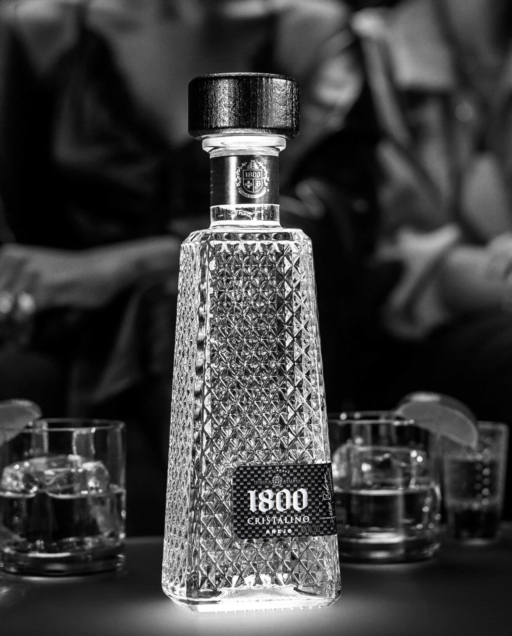 1800 Cristalino Añejo Tequila bottle in crystal design, surrounded by glasses of tequila on a dimly lit table.
