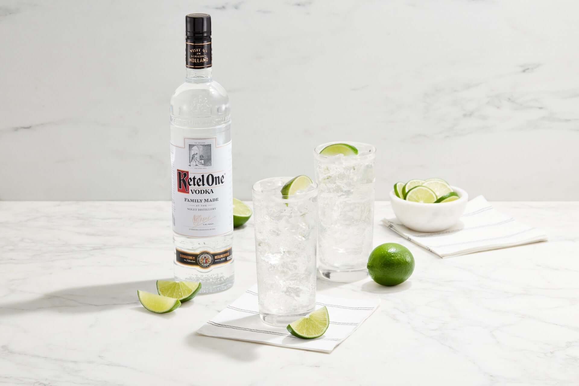 Ketel One Vodka bottle with cocktails and limes on a marble countertop, showcasing freshness and crisp taste.