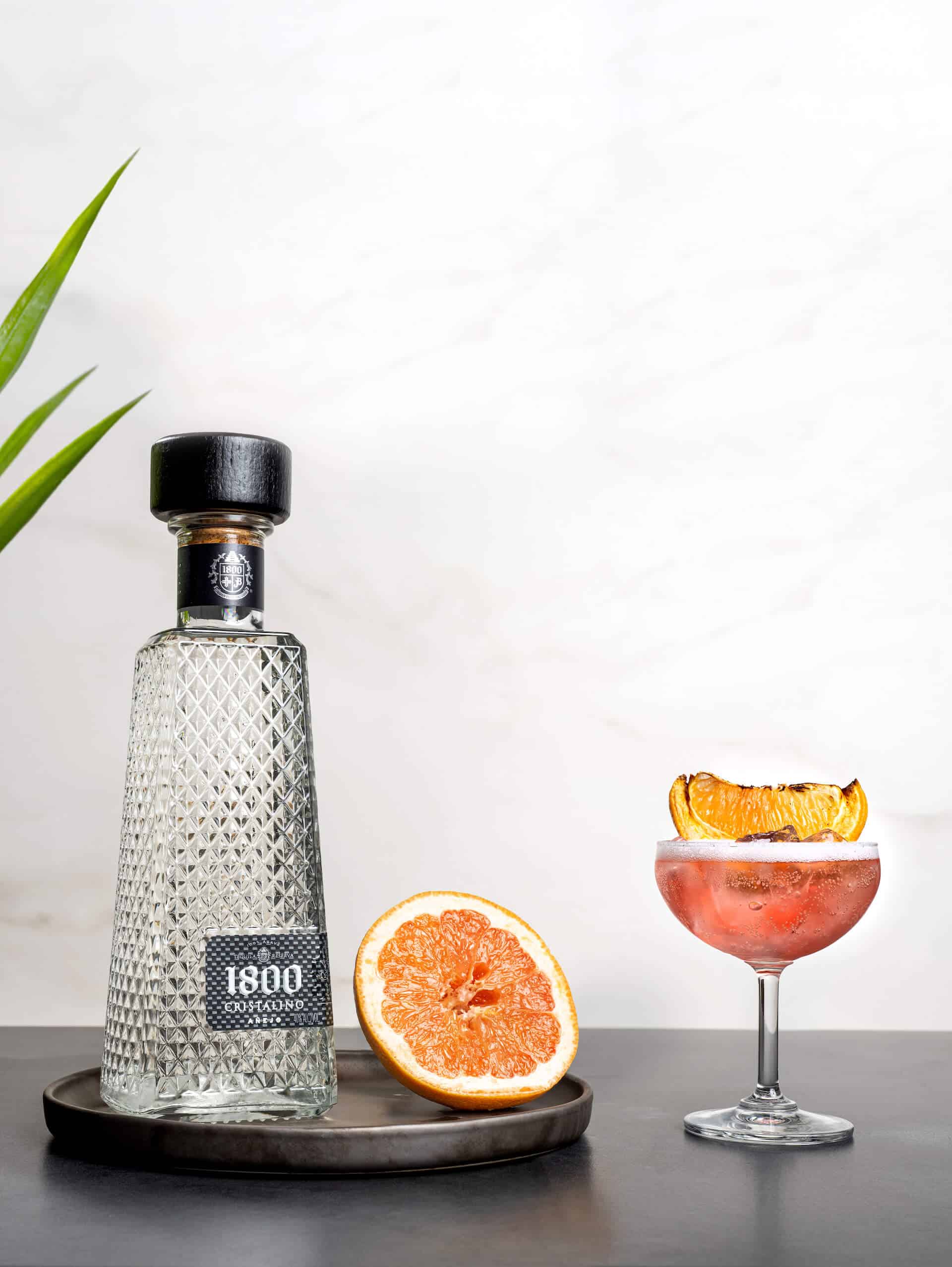 1800 Cristalino Añejo tequila bottle beside a cocktail garnished with grapefruit on a dark surface.