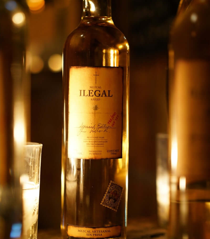 Bottle of Ilegal Mezcal Añejo showcasing its rich, golden hue and artisanal label, set against a soft-lit background.