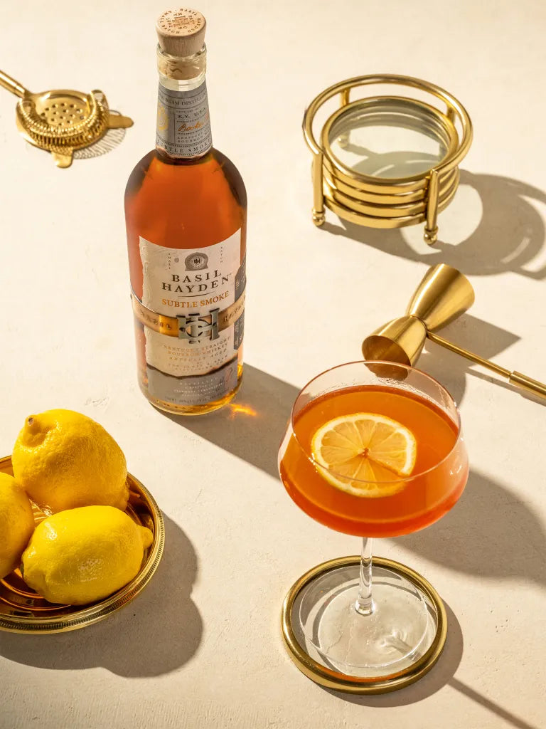 Basil Hayden Subtle Smoke bourbon bottle with cocktail and lemons on a stylish table setting.