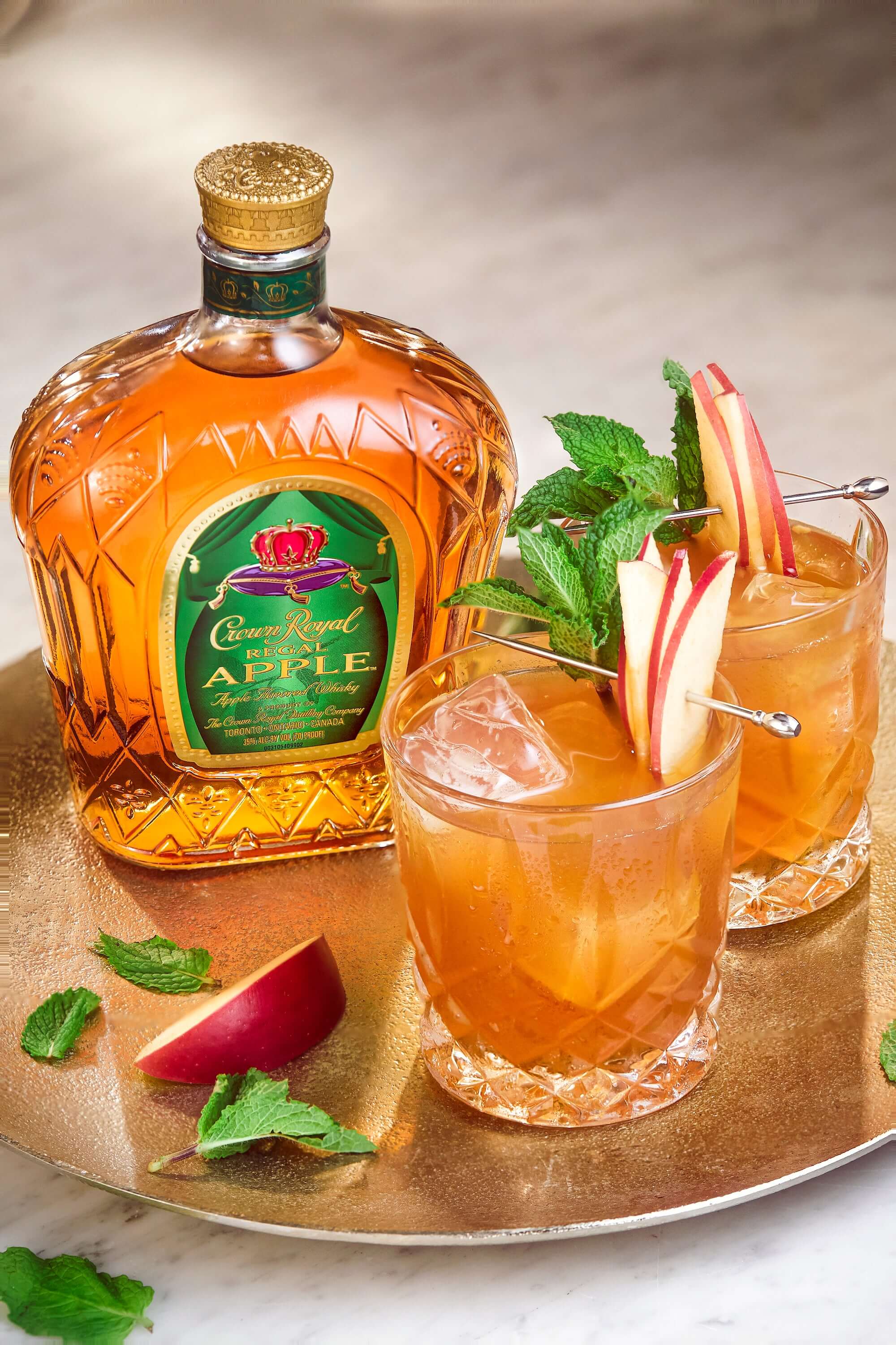 Crown Royal Apple whisky with cocktails garnished with apple slices and mint on a decorative tray.