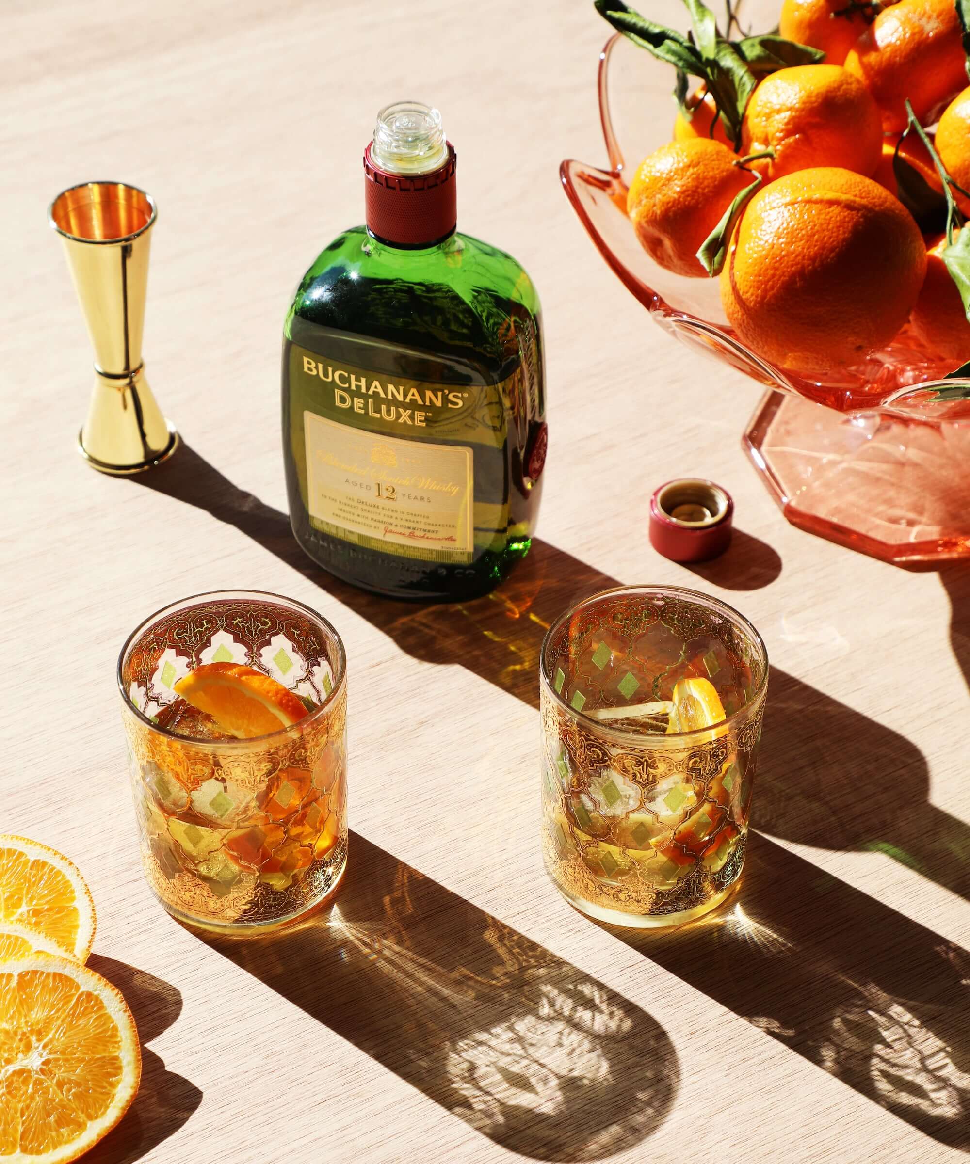Buchanan's Deluxe 12 Year whisky bottle with cocktails and fresh oranges on a wooden table, showcasing its luxury and flavor.