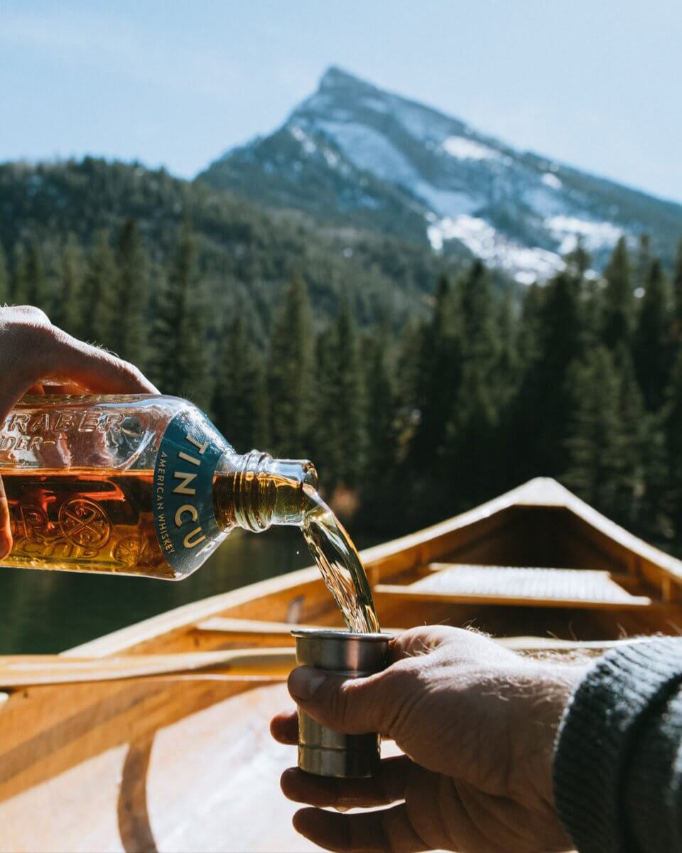 Pouring TINCUP American Whiskey from a bottle into a cup on a scenic lake with mountains in the background.